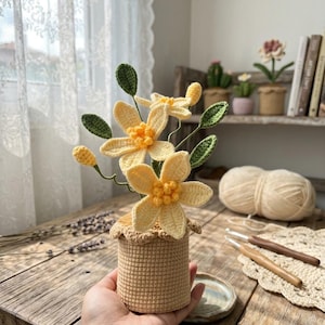 May include: A handmade crochet flower arrangement featuring yellow flowers with green leaves in a small, tan crochet pot. The arrangement is held in a hand, with a wooden table and crochet supplies in the background.