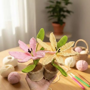 May include: Crocheted lily flowers in pink and cream, each in a small brown pot. The flowers have green leaves and black stamens. The image shows the flowers on a wooden surface with yarn and crochet hooks.