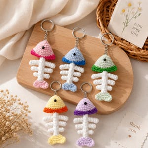 May include: Five crochet fish skeleton keychains with metal rings. Each features a white spine and tail with a colored head in pink, blue, green, yellow, or purple. Each head has a small black bead eye. The keychains are arranged on a wooden board.