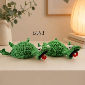 May include: Two green crocheted turtle figures with textured shells, white eyes with black pupils, and open mouths featuring small red tongues. The text Style 1 is positioned above the figures. The items are crafted from green yarn.