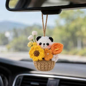 May include: A crocheted car hanging ornament featuring a small white and black panda head nestled in a tan basket filled with yellow sunflowers, orange calla lilies, and small white flowers, suspended by a brown string from a rearview mirror.