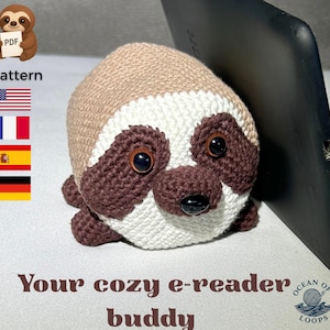 Könnte beinhalten: Ein gehäkeltes Faultier als E-Reader-Buddy in Braun-, Weiß- und Dunkelbrauntönen, mit großen, dunklen Augen und einer schwarzen Nase. Der Text "Your cozy e-reader buddy" ist unter dem Faultier zu sehen. Das Wort "pattern" ist ebenfalls sichtbar.