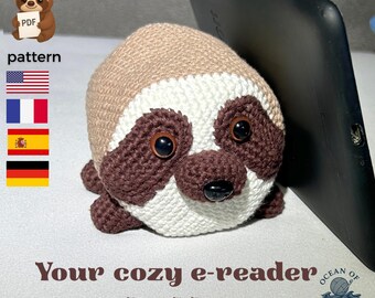 May include: A crocheted sloth e-reader buddy in shades of brown, white, and dark brown, with large, dark eyes and a black nose. The text "Your cozy e-reader buddy" is displayed below the sloth. The word "pattern" is also visible.
