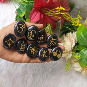 May include: Ten black, oval stones, each featuring a gold symbol. The stones are held in a hand, with red and white flowers in the background. The symbols are diverse in their design.