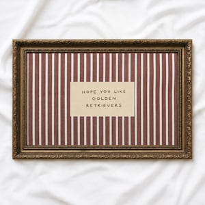 Puede incluir: Un marco de fotos rectangular con un acabado dorado antiguo ornamentado que contiene un fondo de tela con un patrón de rayas verticales granates y blancas. Una tarjeta rectangular de color crema está centrada sobre la tela con el texto HOPE YOU LIKE GOLDEN RETRIEVERS.
