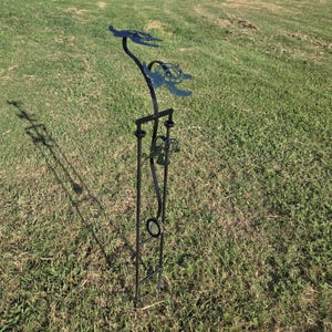May include: A black metal garden stake with a turtle design. The stake features two turtle silhouettes at the top and a decorative element with a ring and other shapes. The stake is placed in green grass.