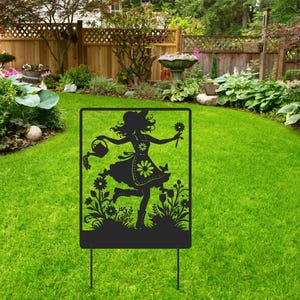 May include: A black metal garden stake featuring a silhouette of a woman in a floral dress, holding a basket and a flower. The decorative stake is set in a green lawn, with a garden and trees in the background.