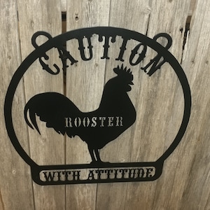 May include: Black metal sign featuring a rooster silhouette within a circular frame. The sign reads "CAUTION ROOSTER WITH ATTITUDE" in capital letters. The sign has two small loops at the top for hanging and is set against a weathered wooden background.