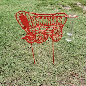 May include: Red metal wagon silhouette with the word "SOONERS" cut out. The wagon has a rain gauge attached to the side.