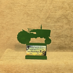 May include: A green metal tractor silhouette with a rectangular base. The tractor has a detailed design, including wheels and a driver's seat. A business card is attached to the base, displaying the company name, location, and website.
