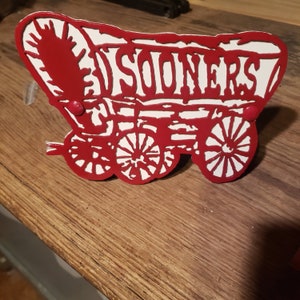 May include: Red metal wagon cutout with white lettering that reads "Sooners". The wagon has two wheels and a handle.