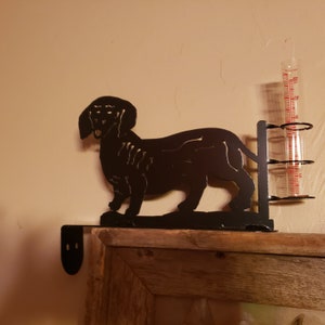 May include: Black metal dachshund silhouette rain gauge holder with a clear glass rain gauge. The holder is mounted on a wooden shelf.