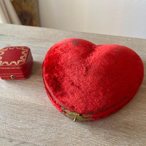 May include: A red heart-shaped jewelry box with a gold clasp next to a small red square box with gold trim. The heart-shaped box is made of a plush material. The square box has a gold border.