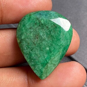 Op de afbeelding: Een peervormige, gefacetteerde smaragd edelsteen. De edelsteen heeft een diepe, rijke groene kleur met zichtbare insluitsels en een gepolijst oppervlak. De steen wordt tussen de vingers gehouden, wat de grootte en vorm laat zien.