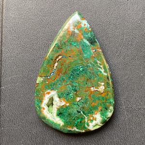 Puede incluir: Una gema en forma de lágrima con una vibrante mezcla de tonos verdes, naranjas y blancos. La superficie pulida revela patrones intrincados y una pequeña cavidad, mostrando la belleza natural de la piedra. La gema está sobre un fondo oscuro.