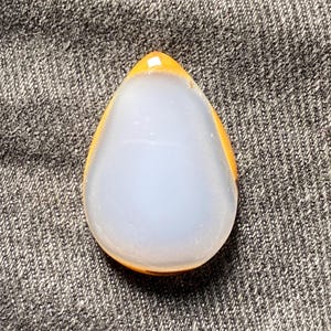 May include: A teardrop-shaped pendant with a light blue center and a golden-orange border. The polished stone has a smooth, glossy surface. The pendant is likely for jewelry making or as a decorative element.