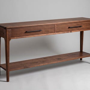 May include: A rectangular wooden console table featuring two drawers with a vertical ribbed texture on the front panels. The table has a smooth top surface, four tapered legs, and a lower shelf. Each drawer is fitted with a thin black handle.