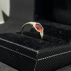 Peut inclure: Bague en argent avec une pierre rouge rectangulaire et facettée. Le bijou est présenté dans un écrin noir avec un intérieur en velours. La pierre précieuse affiche une couleur rouge vif et intense, idéale pour un cadeau.