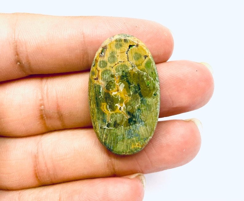 May include: An oval gemstone featuring a distinctive pattern of green, yellow, and brown tones. The stone's surface has a textured appearance with a blend of colours, creating a natural aesthetic. The gemstone is held in a hand.