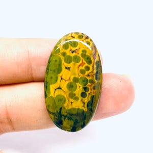 May include: An oval gemstone featuring a distinctive pattern of green and yellow spots. The polished stone has a glossy finish, and the colours blend to create a striking visual effect. The gemstone is held in a person's hand.