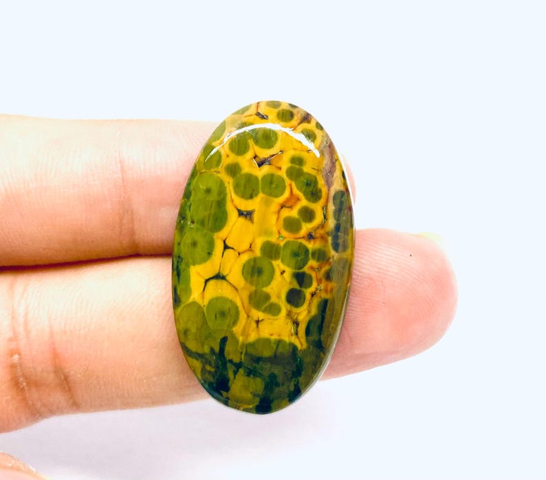 May include: An oval gemstone featuring a distinctive pattern of green and yellow spots. The polished stone has a glossy finish, and the colours blend to create a striking visual effect. The gemstone is held in a person's hand.