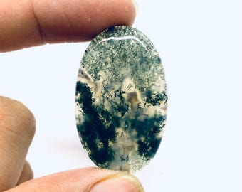 May include: An oval moss agate gemstone. The stone displays a translucent base with detailed dark green and white inclusions, forming a natural pattern. The polished surface reflects light, accentuating the intricate designs. The stone is held between fingers.