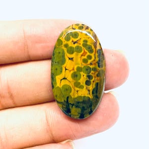 May include: An oval gemstone featuring a distinctive pattern of yellow and green spots. The polished surface reflects light, showcasing the natural colour and texture variations. The gemstone is held in a hand.