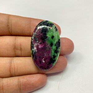 Könnte beinhalten: Ein ovaler Edelstein mit polierter Oberfläche. Der Stein weist eine Mischung aus Farben auf, darunter tiefes Rot, Grün und Schwarz. Die roten und grünen Farbtöne dominieren, mit schwarzen Sprenkeln. Der Edelstein wird in einer Hand gehalten.