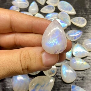 May include: A collection of polished moonstone gemstones. The stones are pear-shaped, with a milky white base and iridescent blue and purple flashes. One stone is held in a hand, showcasing its luster. The background features more moonstones.