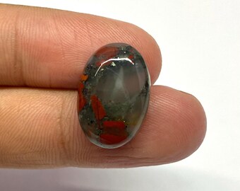 May include: Oval gemstone with a polished surface. The stone displays a mix of red, grey, and green hues, with a translucent appearance. The gemstone is held between fingers against a white backdrop.