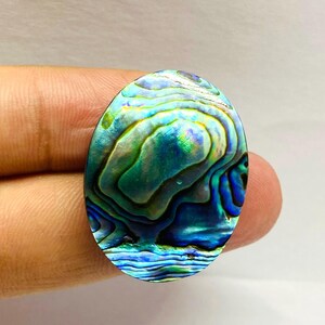 May include: An oval-shaped abalone shell cabochon with iridescent blue, green, and teal swirls. The polished surface reflects light, showcasing the natural patterns and colors of the shell. The gemstone is held by a person's fingers.