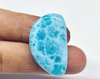 May include: A polished, teardrop-shaped gemstone with a vibrant blue hue and a unique, cloud-like pattern. The stone's surface is smooth and glossy, reflecting light. The gemstone is held by a person's fingers.
