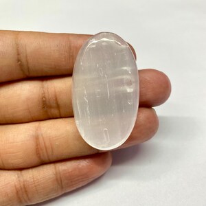 Könnte beinhalten: Ein durchscheinender, oval geformter Selenitkristall. Der Stein hat eine blass cremeweiße Farbe und eine glatte, polierte Oberfläche. Der Kristall wird in einer Hand vor weißem Hintergrund gehalten.