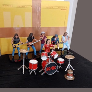 Könnte beinhalten: Ein Set aus fünf Miniatur-Musikerfiguren und einem Schlagzeug, dessen Bassdrum das AC/DC-Logo trägt. Die Bandmitglieder sind mit Gitarren, einem Mikrofon und Trommeln dargestellt. Eine kleine bronzefarbene Glocke steht vor dem Schlagzeugset.