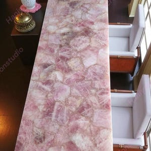May include: A rectangular table with a pink, mosaic-like surface made of rose quartz. The table is surrounded by two white armchairs with wooden frames. A small table with a decorative gold bell and a white cup is visible.