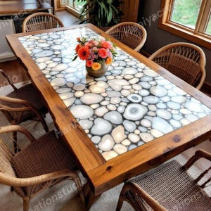 May include: A dining table with a distinctive design. The tabletop is a mosaic of white and grey agate stones set in clear resin. The table has a dark wood frame and is surrounded by woven chairs. A vase of flowers sits in the centre.