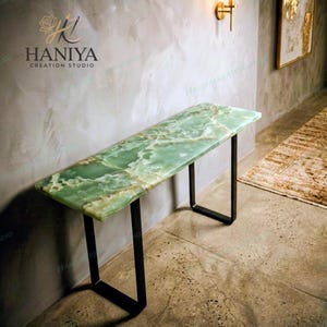 May include: A rectangular console table featuring a polished green onyx stone top with natural white veining and a smooth finish. The table is supported by two black metal U-shaped legs that provide a stable base for the heavy stone surface.