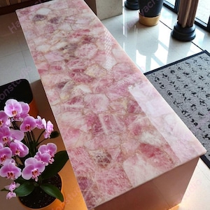 May include: A rectangular coffee table with a rose quartz top. The table's surface is a mosaic of pink and white stones, creating a unique pattern. The base of the table is a solid, light-colored material. The table is in a well-lit room.