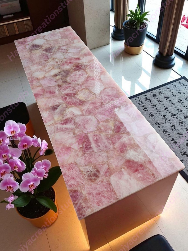 May include: A rectangular coffee table with a rose quartz top. The table's surface is a mosaic of pink and white stones, creating a unique pattern. The base of the table is a solid, light-colored material. The table is in a well-lit room.