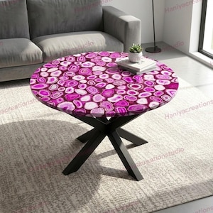 May include: A circular coffee table featuring a pink agate stone top. The table has a dark, cross-shaped base. A small plant and books are on the table. The table is in a living room setting.