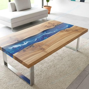 Op de afbeelding: Een rechthoekige houten salontafel met een blauw hars rivierontwerp. De tafel heeft een natuurlijke houtnerf afwerking en staat op zilveren metalen poten. De blauwe hars heeft witte accenten.