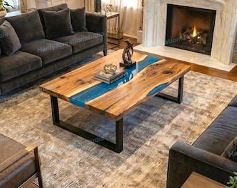 May include: A rectangular wooden coffee table with a blue resin river design. The table has a natural wood grain finish and sits on black metal legs. The table is in a living room setting with a fireplace and sofa.