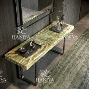 May include: A rectangular console table featuring a light green onyx stone top with natural banded patterns and a thick, textured edge. The table is supported by two dark metal U-shaped legs. A dark rectangular tray holds a small bowl and keys.