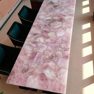 May include: A rectangular table with a pink rose quartz top. The table's surface is composed of irregular, interlocking pieces of pink and white stone. The table is supported by a black base, and is surrounded by dark green chairs.