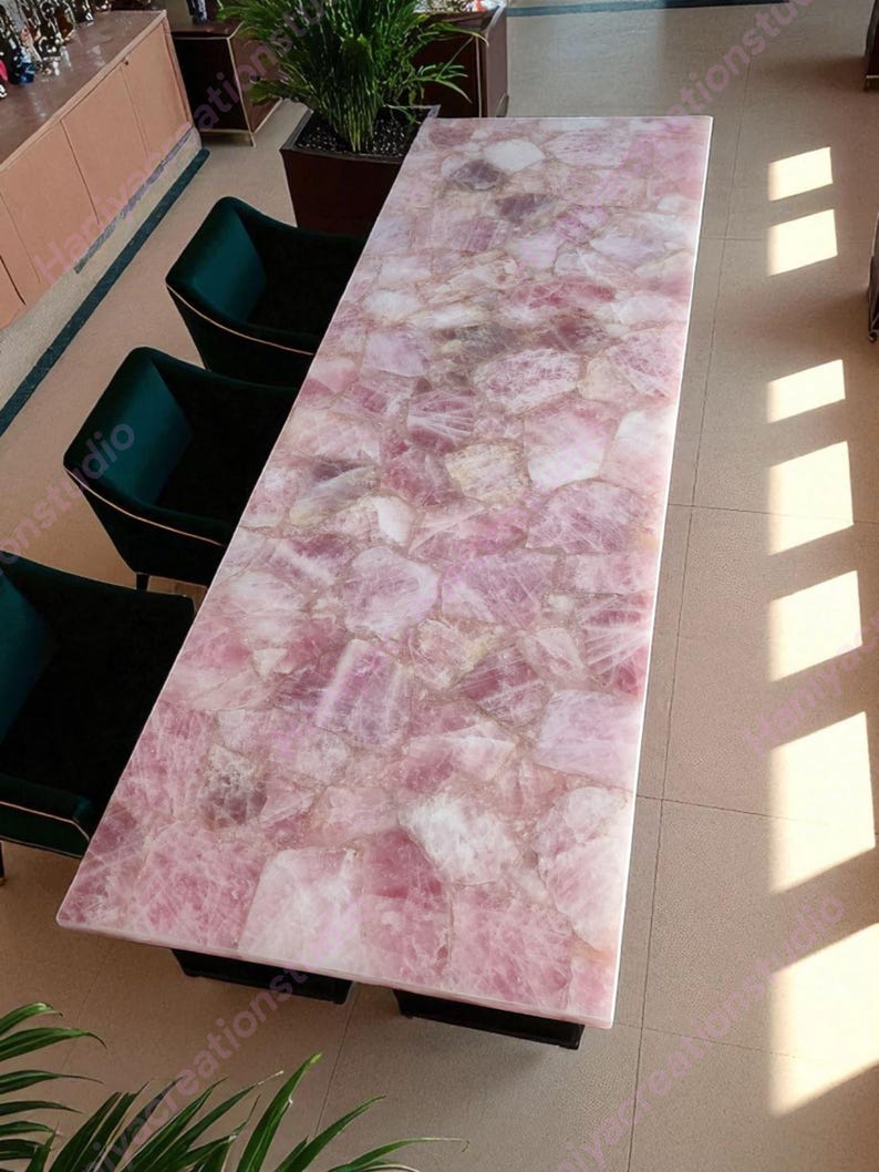 May include: A rectangular table with a pink rose quartz top. The table's surface is composed of irregular, interlocking pieces of pink and white stone. The table is supported by a black base, and is surrounded by dark green chairs.