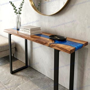 May include: A wooden console table with a live edge design and a vibrant blue resin river. The table has black metal legs and is topped with books and a black tray. The table is approximately 48 inches long.