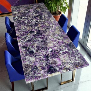 Puede incluir: Una mesa de comedor rectangular de amatista con una superficie pulida, sostenida por patas de metal color oro. La mesa está rodeada por cuatro sillas azules. Una planta en maceta y una obra de arte son visibles en el fondo.