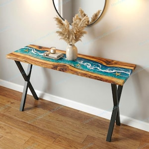 Könnte beinhalten: Holzkonsole mit einem blauen Epoxidharz-Flussdesign. Der Tisch hat schwarze Metallbeine und ist mit Seesternen verziert. Eine Vase mit Trockenblumen und ein Spiegel befinden sich im Hintergrund.
