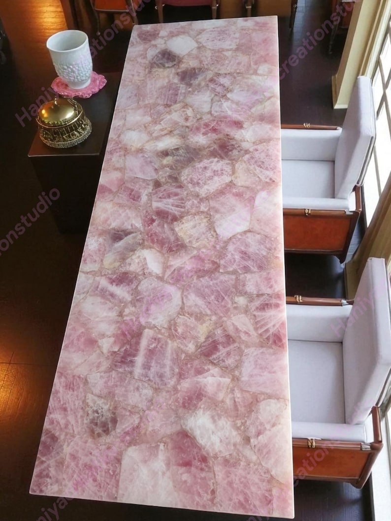May include: A rectangular table with a pink, mosaic-like surface made of rose quartz. The table is surrounded by two white armchairs with wooden frames. A small table with a decorative gold bell and a white cup is visible.
