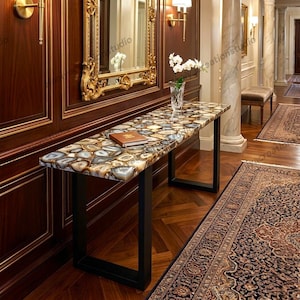 May include: A rectangular console table with a mosaic agate stone top and black metal legs. The table is in a hallway with a dark wood paneled wall, a large mirror, and a patterned rug. A book and a vase of flowers are on the table.